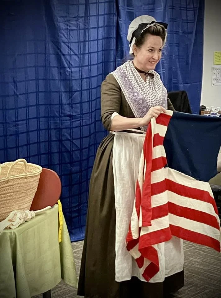 Jen in period attire holds an American flag beside a sewing display.