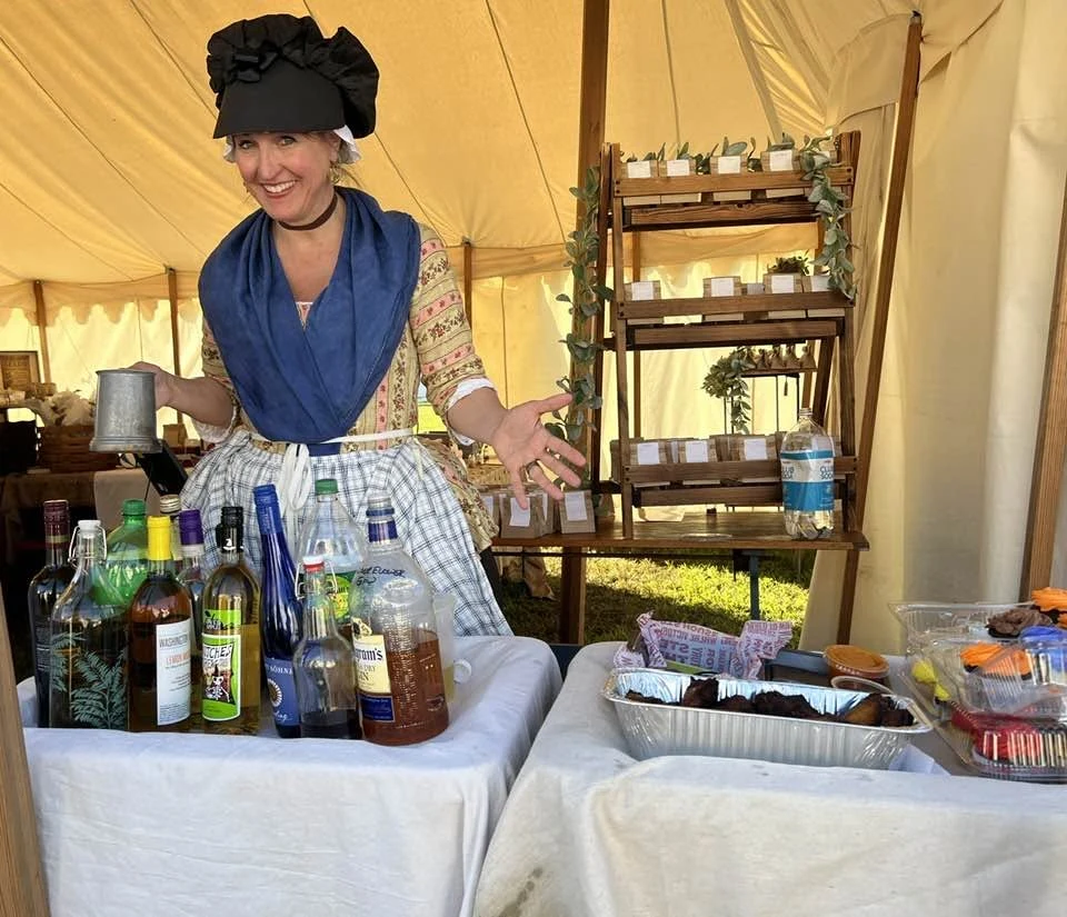 Jen in period attire stands behind a display of bottles, glasses, and historical objects.