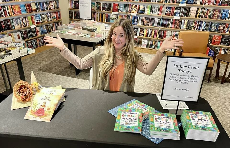 Jen welcomes visitors at a library author-event table with her books.