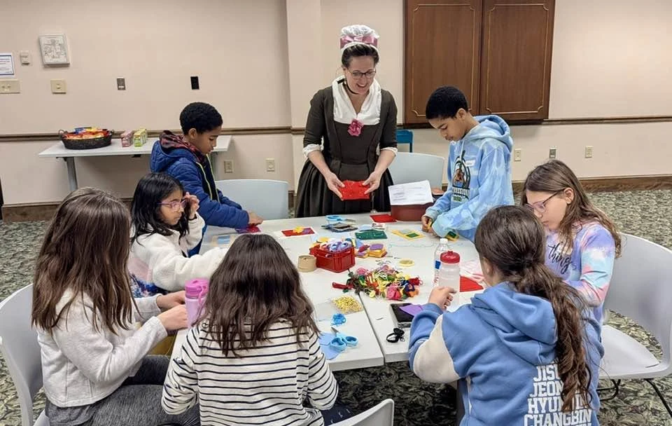 Jen leads a group of children making projects around a workshop table.